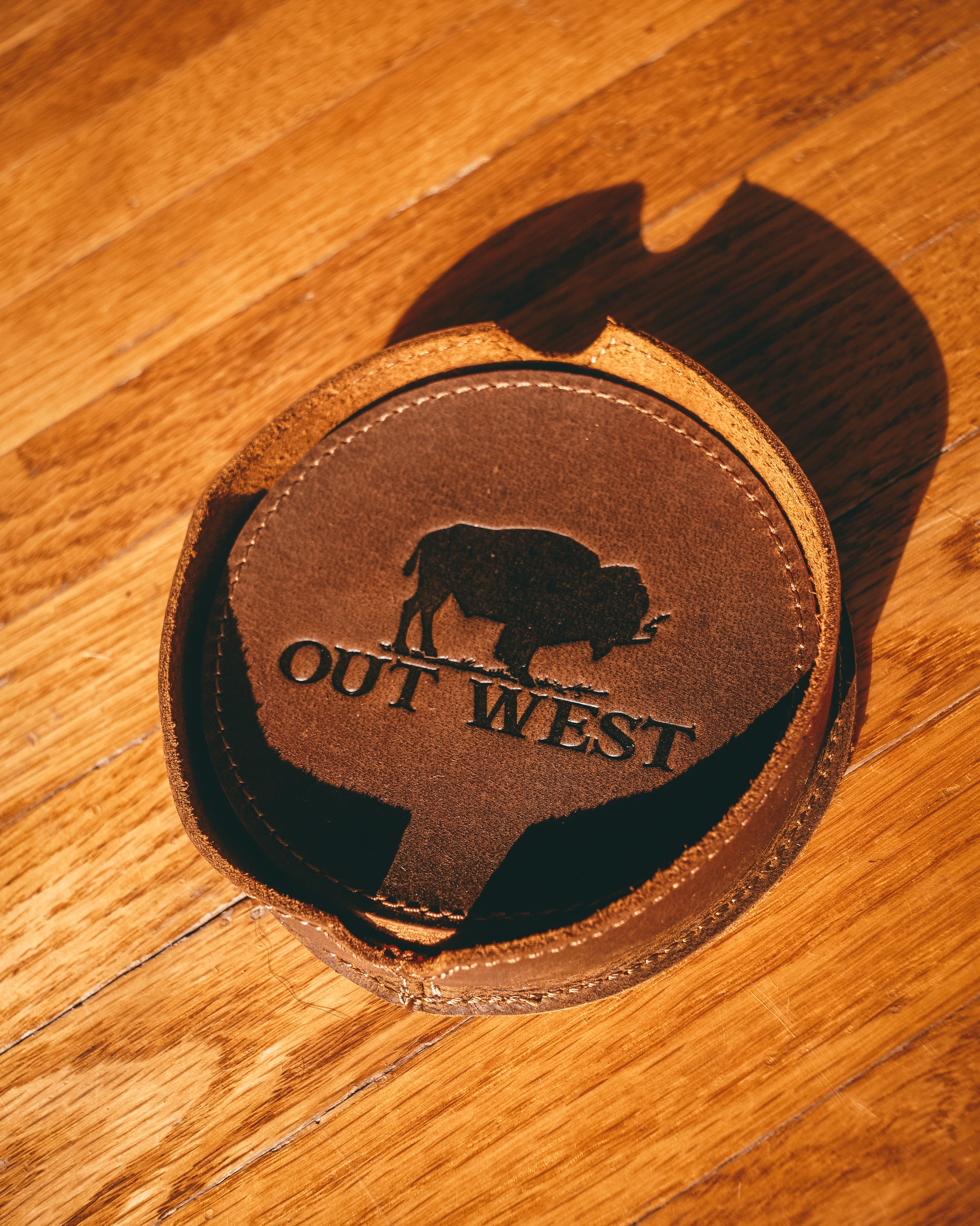 Genuine leather Out West coasters-  Custom designed by Out West and made with genuine leather! Each coaster is branded with the Out West bison logo. Coasters are in a set of 6 + a holder. These coasters will add a true western feel to any home or bar!  Color: Brown  Coaster dimensions: 10cm x 10cm