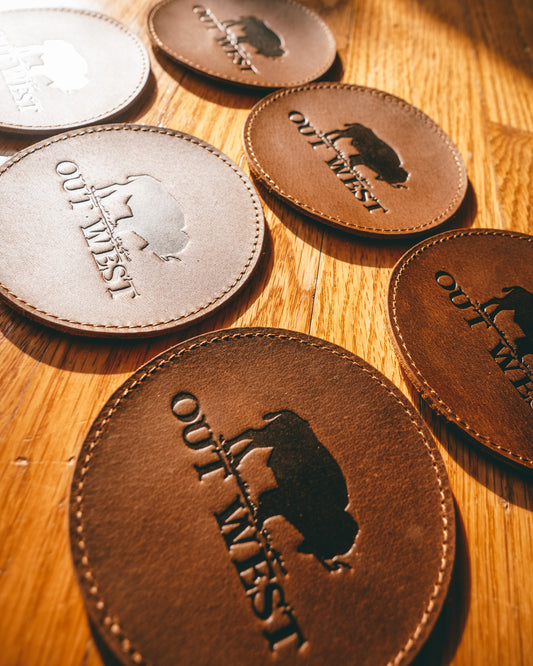 Genuine leather Out West coasters-  Custom designed by Out West and made with genuine leather! Each coaster is branded with the Out West bison logo. Coasters are in a set of 6 + a holder. These coasters will add a true western feel to any home or bar!  Color: Brown  Coaster dimensions: 10cm x 10cm